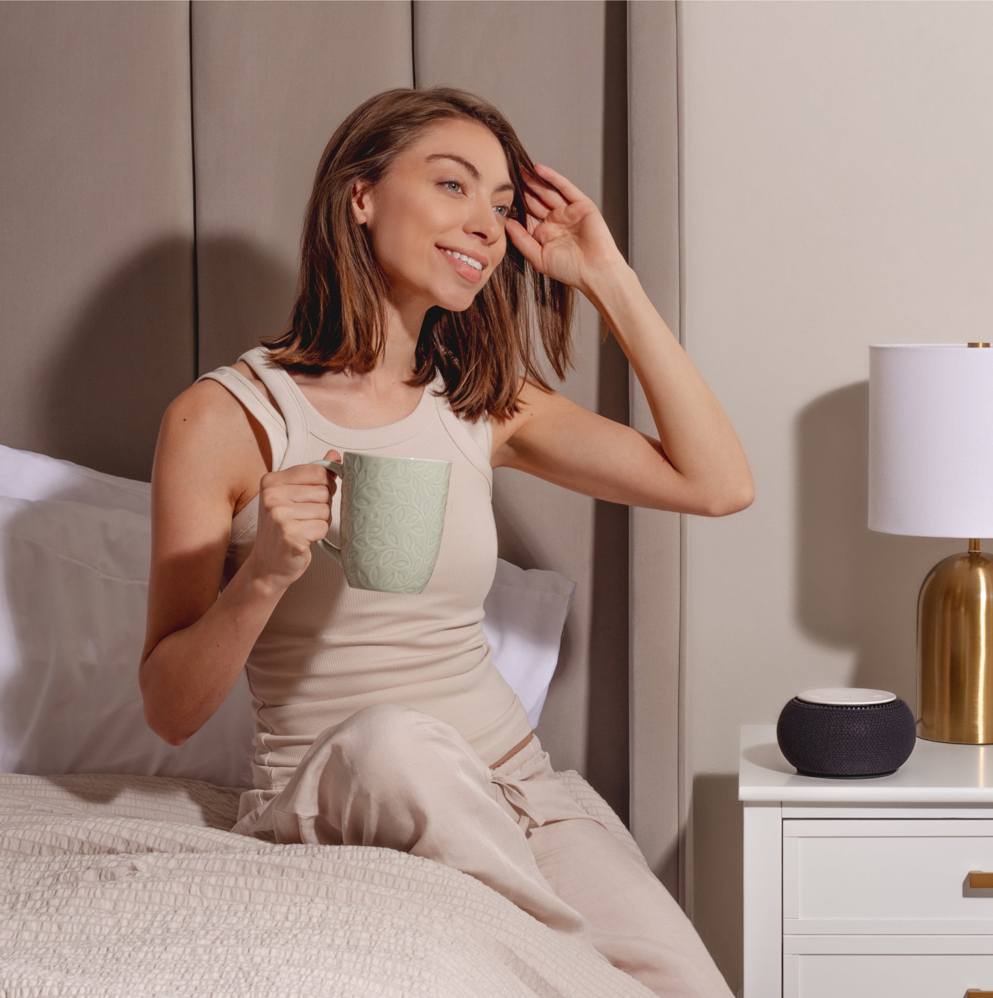Person sitting on a bed, holding a mug and smiling, with a nightstand and lamp beside them.